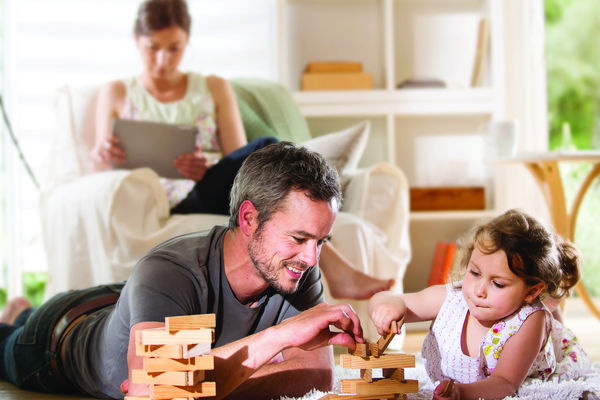 Cheerful family at home, Daddy and his young daughter lying on the wooden floor are playing with a wooden game. At background mom is sitting in an armchair while using a digital tablet