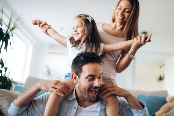 Family girl on dads shoulders, mom behind. livingroom sofa